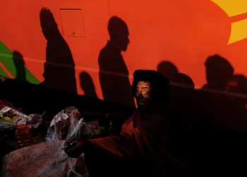 Man sitting in shadow beside a red bus with crowd silhouettes above