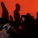 Man sitting in shadow beside a red bus with crowd silhouettes above