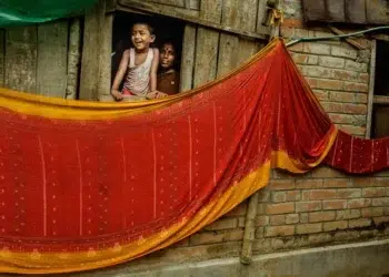 Laughing child and woman peering from window above vibrant red drying saris