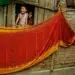 Laughing child and woman peering from window above vibrant red drying saris