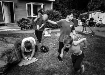 Family in a cluttered yard, child running while woman writes on cardboard, black and white photo