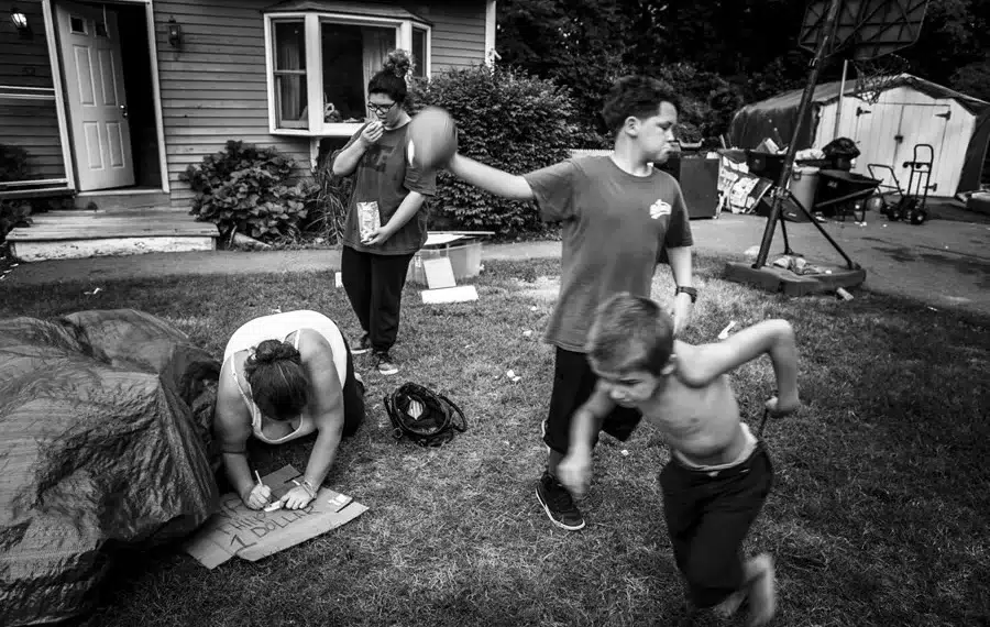 Family in a cluttered yard, child running while woman writes on cardboard, black and white photo