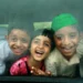 Three laughing children looking through a bus window with joyful expressions