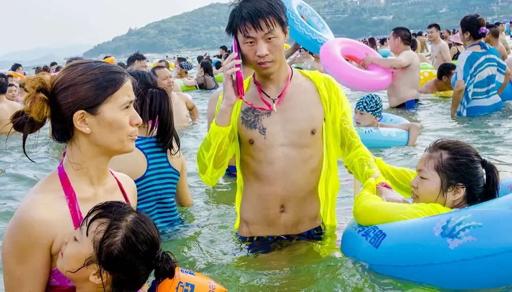 Man on phone in neon jacket amid crowded beach swimmers with colorful floats