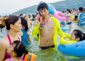 Man on phone in neon jacket amid crowded beach swimmers with colorful floats