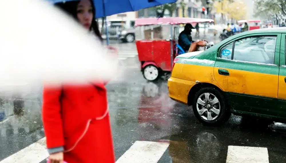 Woman in a red coat with a blue umbrella crossing a rainy city street