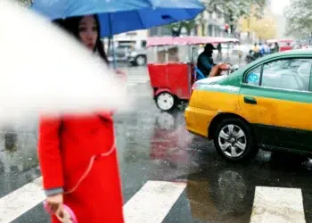 Woman in a red coat with a blue umbrella crossing a rainy city street