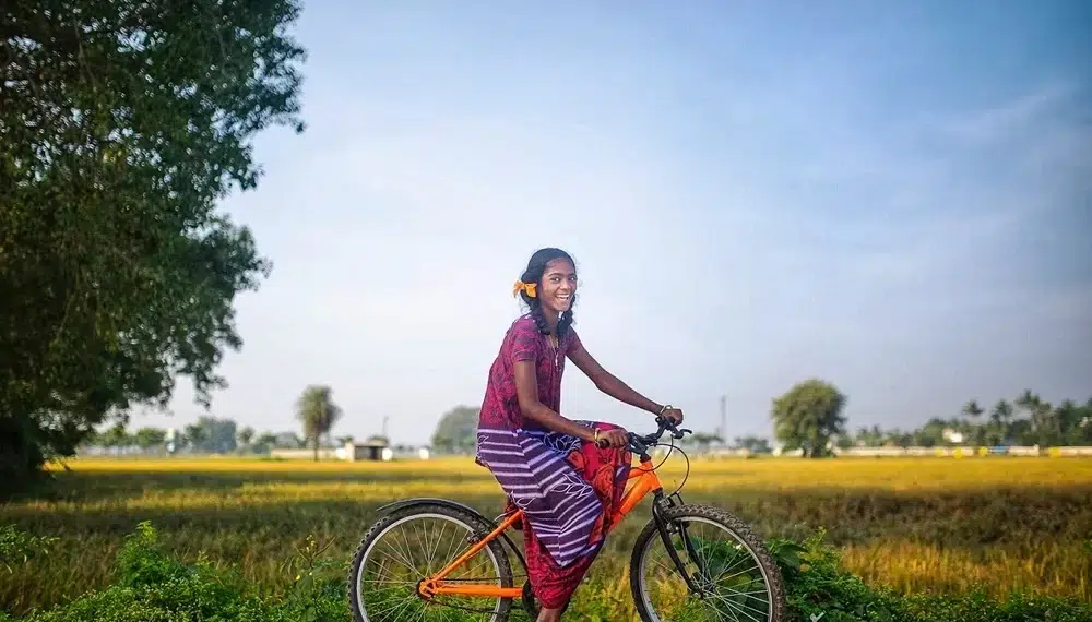 Smiling girl on an orange bicycle beside green fields in a rural setting