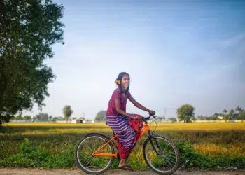 Smiling girl on an orange bicycle beside green fields in a rural setting