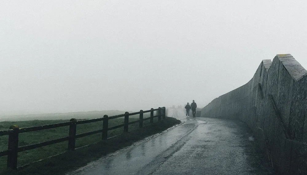 Two figures walking a misty coastal path beside a stone wall, Ireland