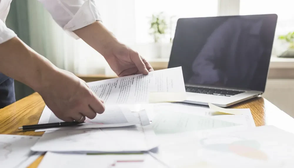 Person reviewing documents at a desk with a laptop, papers, and sticky notes