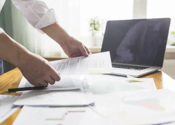 Person reviewing documents at a desk with a laptop, papers, and sticky notes