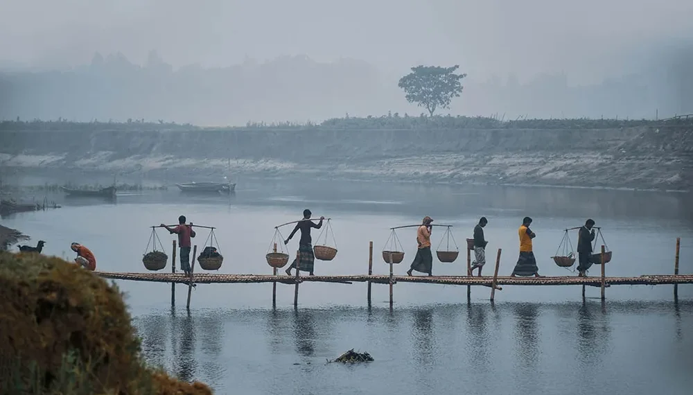 Villagers carrying basket yokes across a bamboo bridge in misty Bangladesh winter