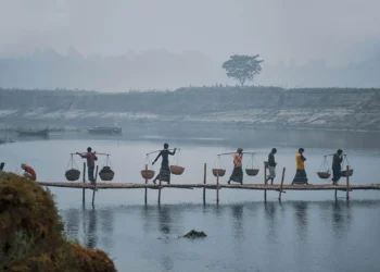 Villagers carrying basket yokes across a bamboo bridge in misty Bangladesh winter