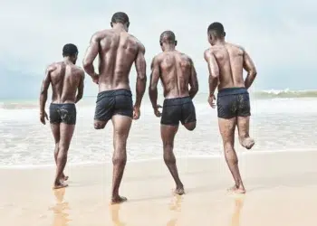 Four men in swim shorts running toward ocean waves on a sandy beach.