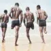 Four men in swim shorts running toward ocean waves on a sandy beach.