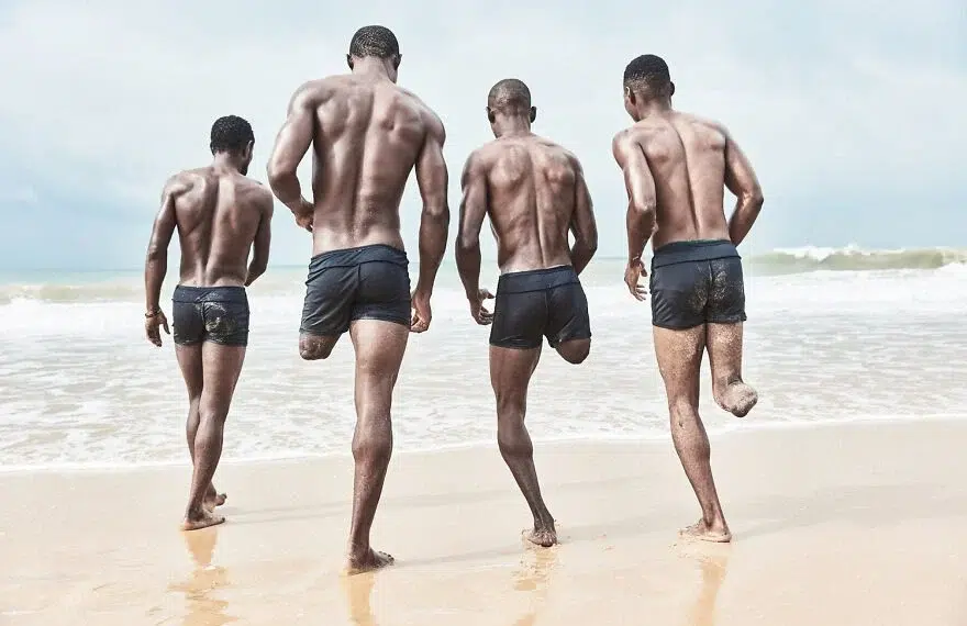 Four men in swim shorts running toward ocean waves on a sandy beach.