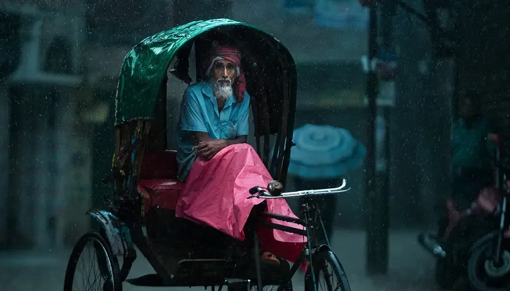Elderly rickshaw driver sheltering under canopy during heavy monsoon rain, Dhaka