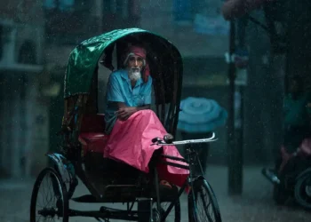 Elderly rickshaw driver sheltering under canopy during heavy monsoon rain, Dhaka