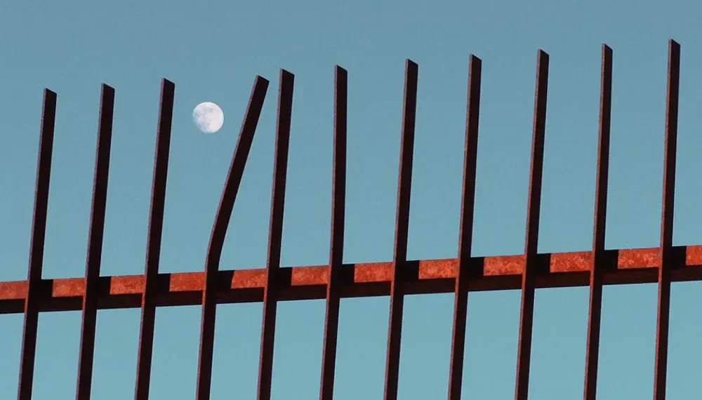 Full moon visible behind rusty iron fence against blue sky