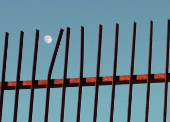 Full moon visible behind rusty iron fence against blue sky