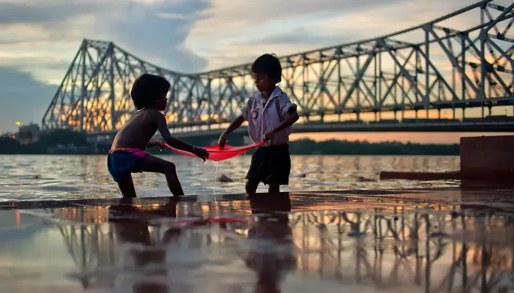 Two children playing with red cloth by Howrah Bridge at dusk
