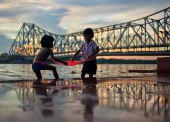 Two children playing with red cloth by Howrah Bridge at dusk