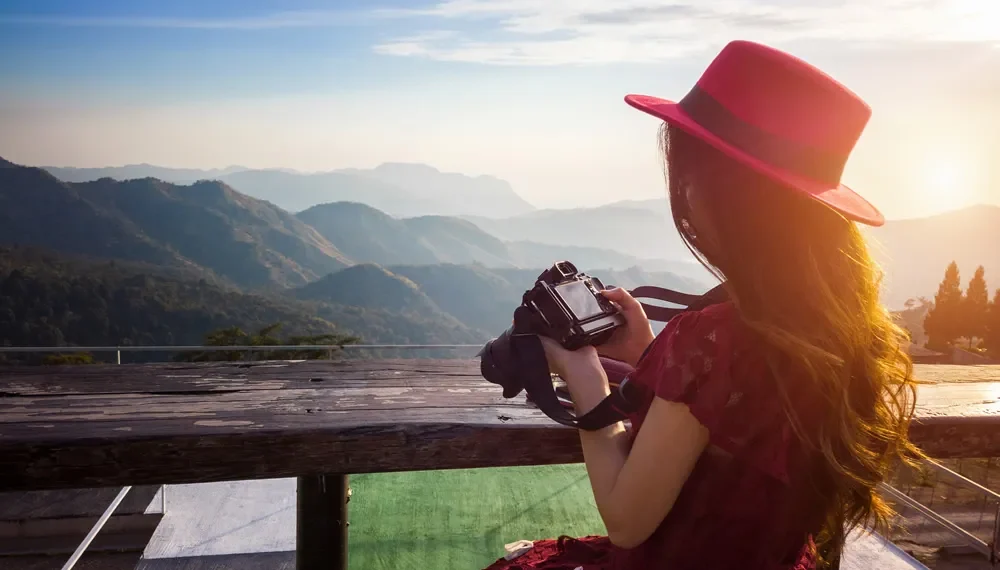 Woman in red hat holding a camera overlooking a mountain landscape at sunset