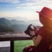 Woman in red hat holding a camera overlooking a mountain landscape at sunset