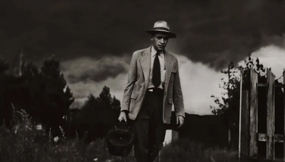 A weathered doctor walks through a field carrying a medical bag, stormy sky above