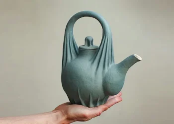 Hand holding a sculptural matte teal ceramic teapot with draped fabric details