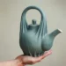 Hand holding a sculptural matte teal ceramic teapot with draped fabric details