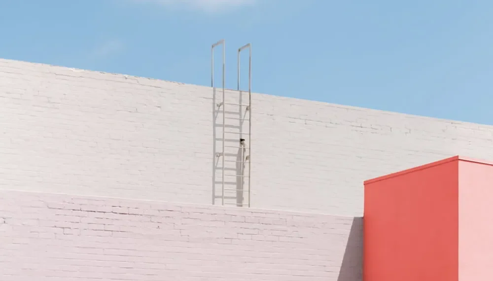 Ladder against a white wall beside a pink building under a blue sky
