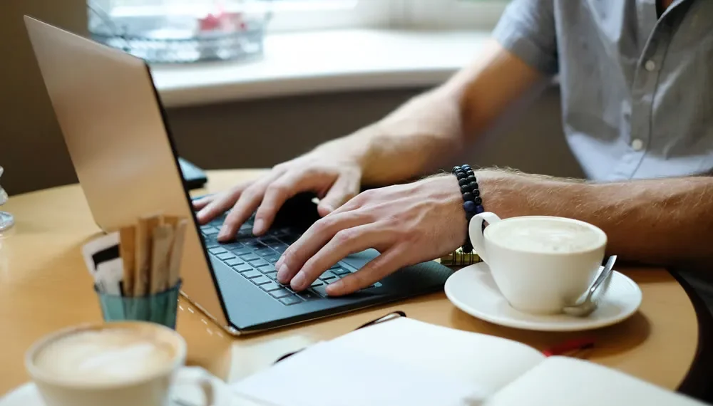 How To Start A Photography Blog: A Step-By-Step Guide 4 Man typing on a laptop at a café table with coffee cups and a notebook