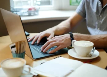 Man typing on a laptop at a café table with coffee cups and a notebook