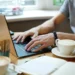 Man typing on a laptop at a café table with coffee cups and a notebook