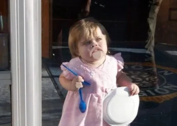 Toddler in pink dress holding a spoon and bowl with food on her face — funny parenting pictures