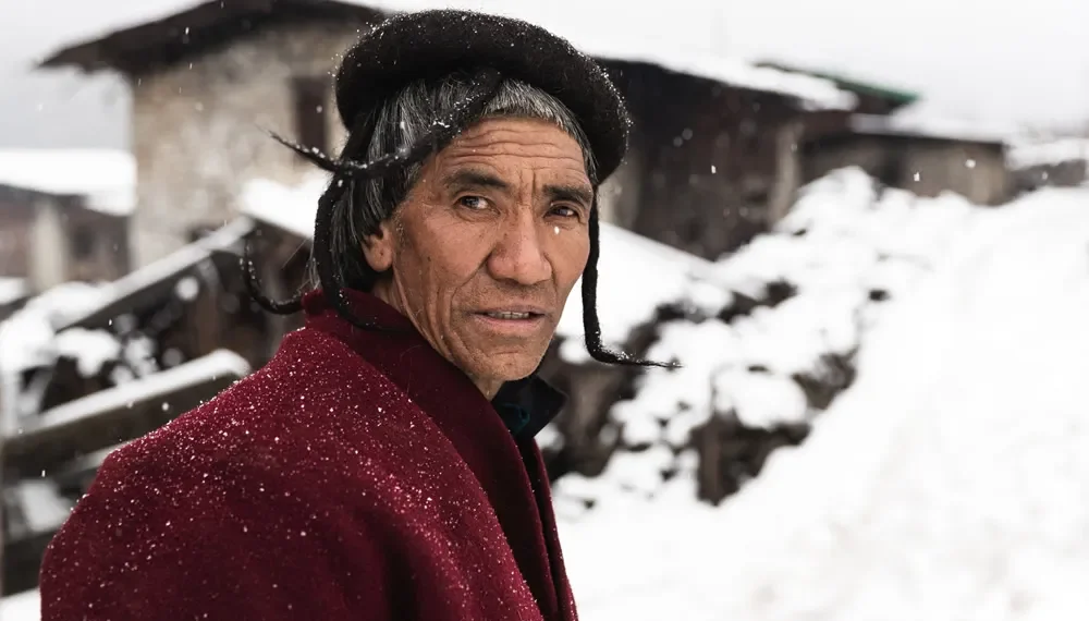 Brokpa man in traditional hat and red coat standing in a snowy Bhutan village