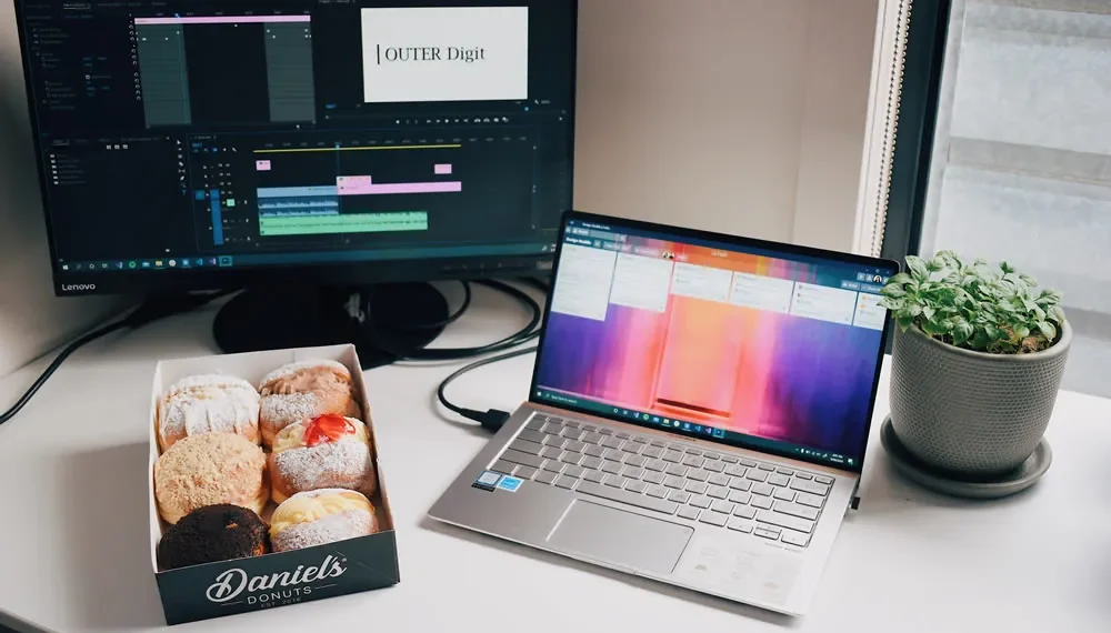 Desk with video editing software on monitor, laptop, box of donuts, and plant