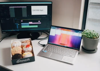 Desk with video editing software on monitor, laptop, box of donuts, and plant