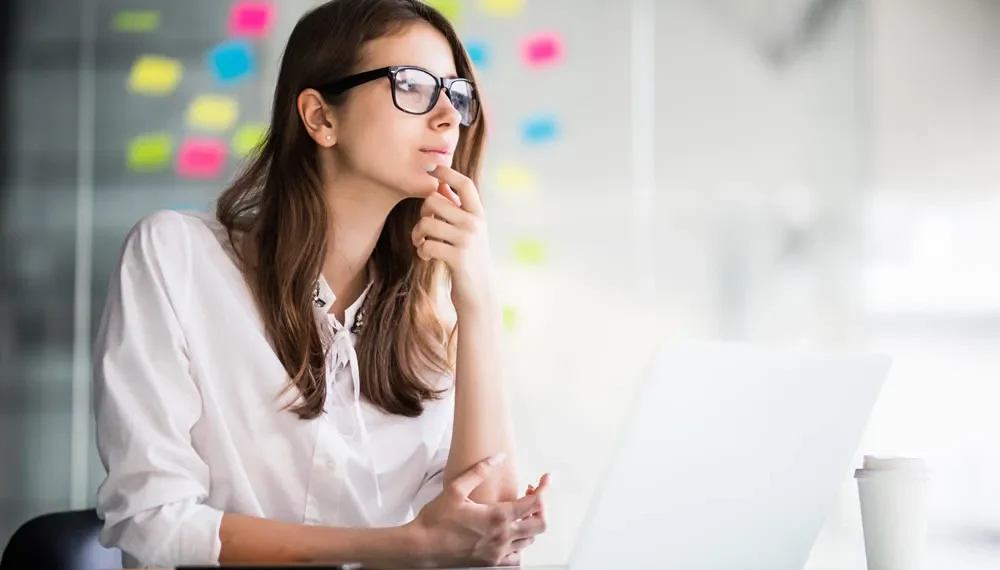 Woman with glasses thinking at a laptop with colorful sticky notes behind