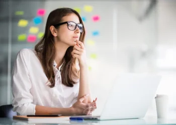 Woman with glasses thinking at a laptop with colorful sticky notes behind