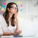 Woman with glasses thinking at a laptop with colorful sticky notes behind