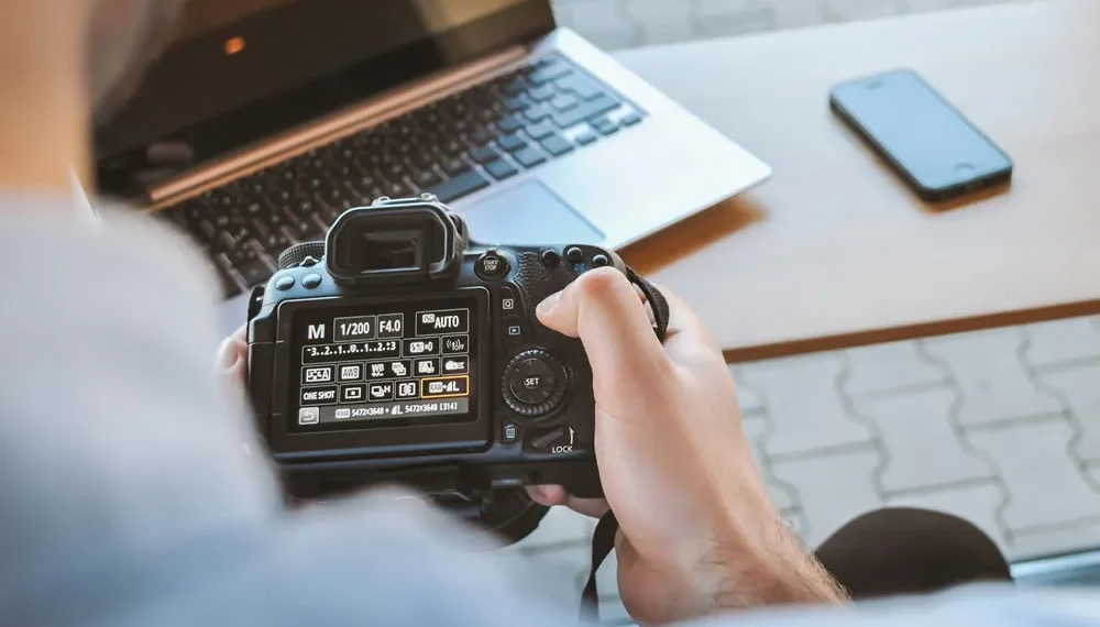 Person holding a DSLR camera showing manual settings screen at a desk