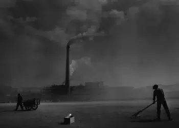 Workers toiling near a smoky factory chimney in a dark b&w industrial scene
