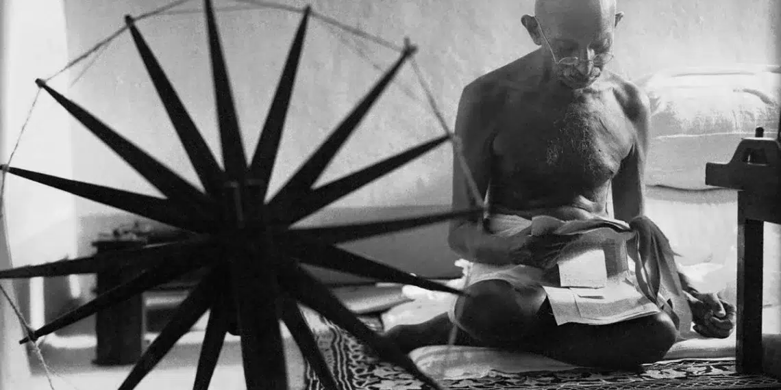 Man seated cross-legged beside a spinning wheel, black and white