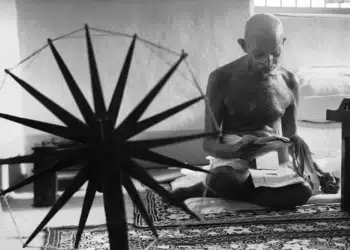 Man seated cross-legged beside a spinning wheel, black and white