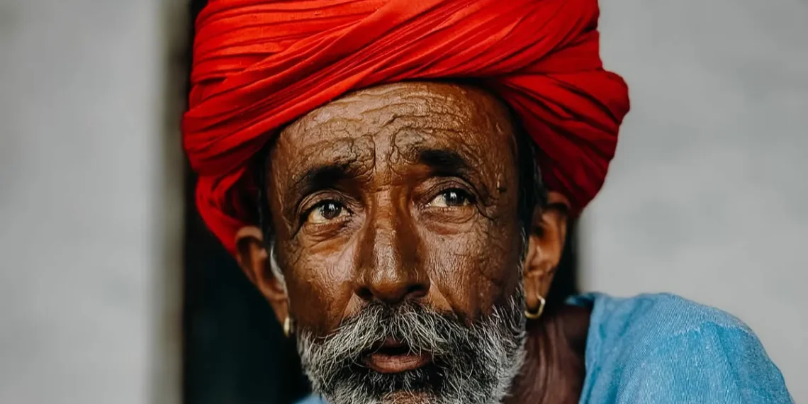 An elderly man with a white beard wearing a vibrant red turban and blue shirt