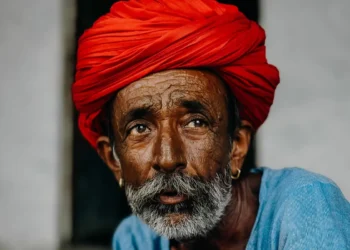 An elderly man with a white beard wearing a vibrant red turban and blue shirt