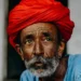 An elderly man with a white beard wearing a vibrant red turban and blue shirt